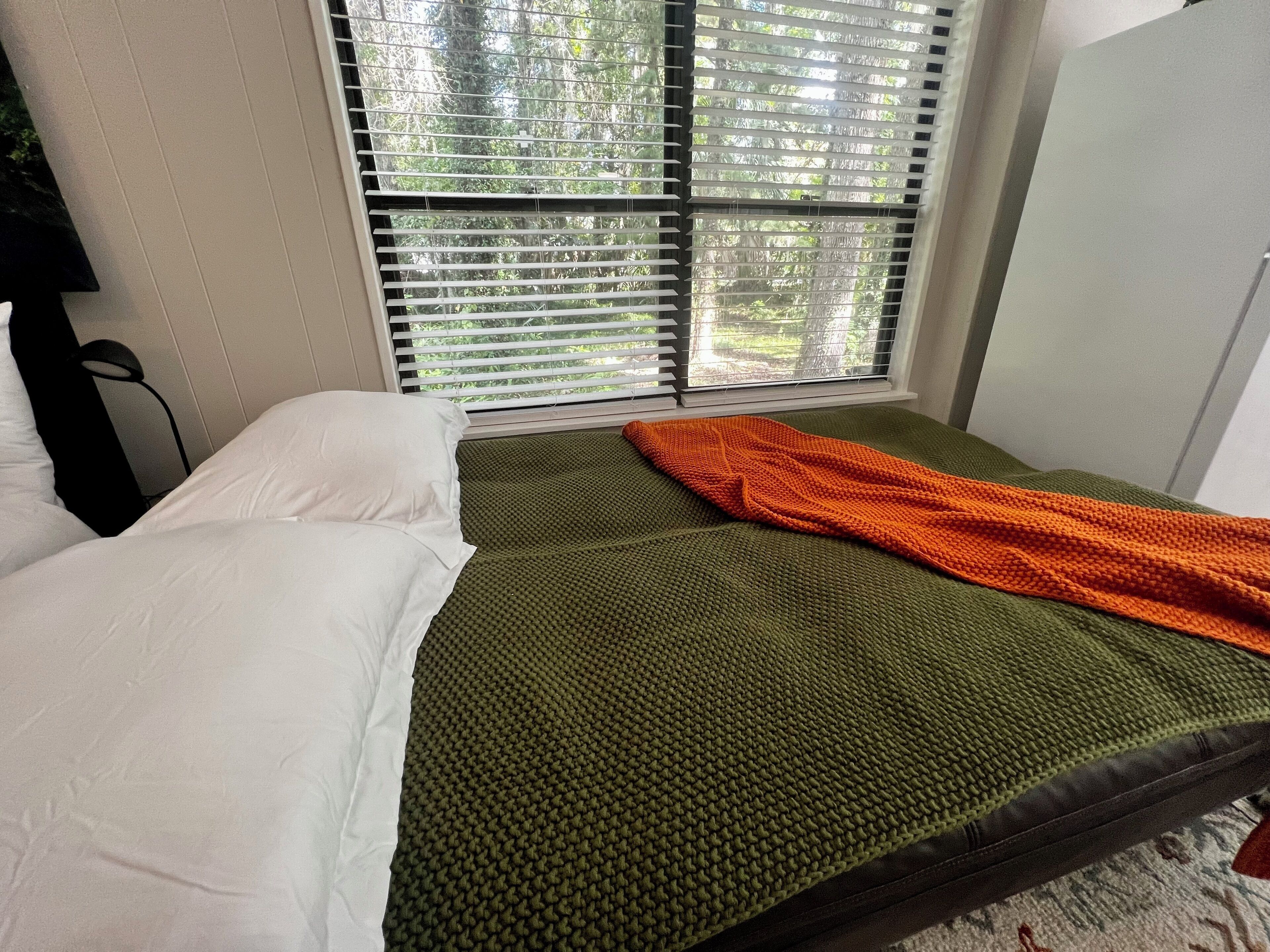 A room with a bed, a window with blinds, and a green textured blanket.