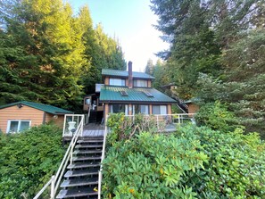 A two-story house with a green roof, surrounded by dense forest. - Waterfront home located in peaceful Bamfield Inlet with private dock. (Bamfield)
