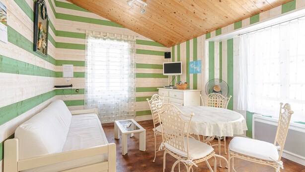 Une pièce avec un plafond en bois, des murs à rayures vertes et blanches, un lit, une table à manger avec des chaises, un téléviseur et une fenêtre avec des rideaux.