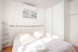 A bedroom with a bed, a wall-mounted air conditioner, a shelf with decorative items, and a painting of a beach scene.