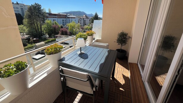 A balcony with a table and chairs, potted plants, and a view of buildings and trees. - Lake - Golden Triangle (Annecy)