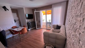 A modern living room with a sofa, a dining area with a table and chairs, a kitchen area with a TV, and a sliding glass door leading to an outdoor patio. - Lake - Golden Triangle (Annecy)