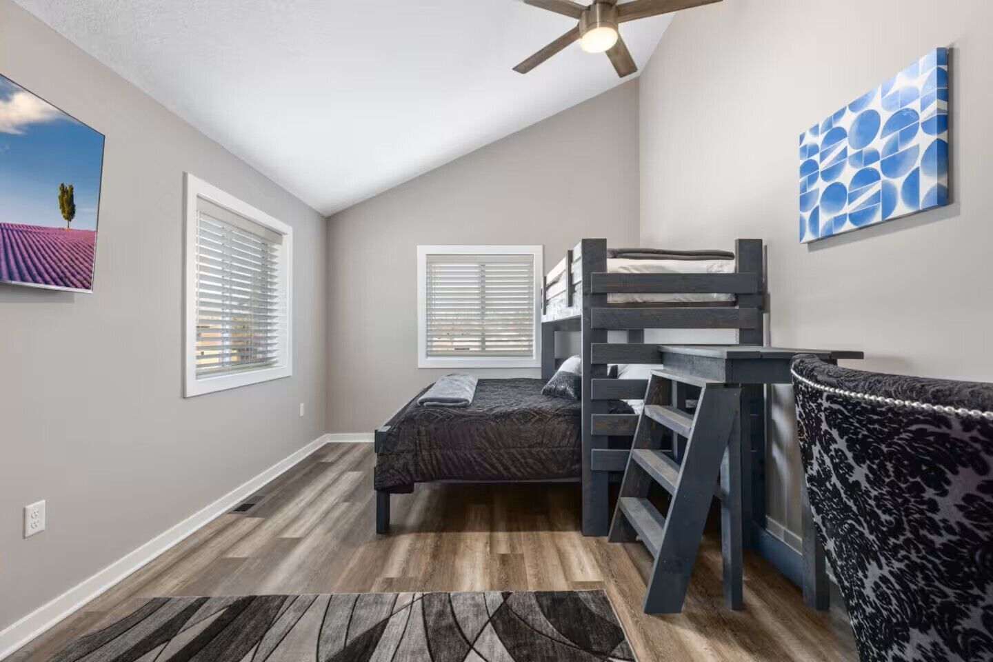 Une chambre avec un lit superposé, un ventilateur de plafond et une grande fenêtre avec des stores.