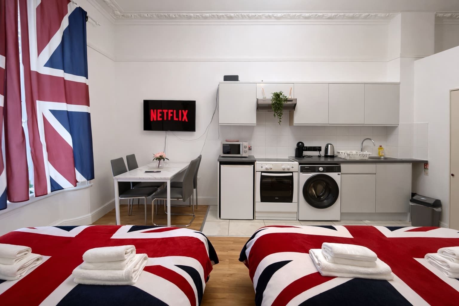Ein kompaktes Hotelzimmer mit einem Bett, das ein Union-Jack-Design zeigt, einer kleinen Küchenzeile mit Waschmaschine, Mikrowelle und einem Fernseher, der 'NETFLIX' anzeigt.