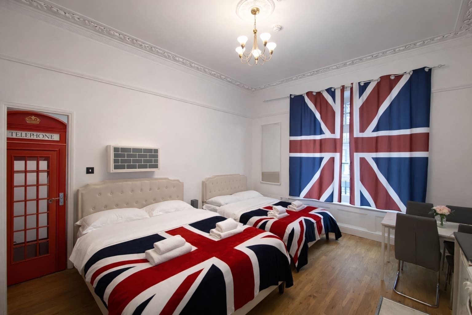 A hotel room with two beds, a telephone, a chandelier, and a large window with a Union Jack curtain.