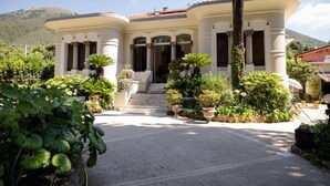 Un edificio a due piani con una facciata curvilinea, un ingresso centrale con gradini e un'area giardino con diverse piante.