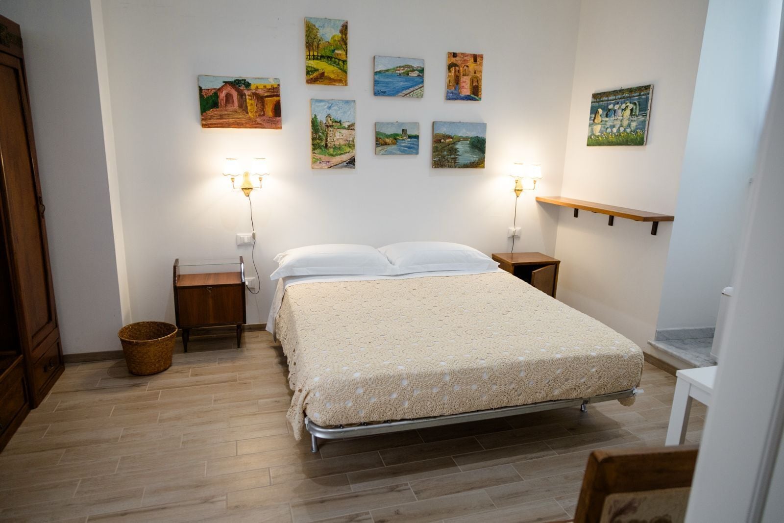 A bedroom with a bed, two bedside tables, a lamp, a wooden floor, and six paintings on the wall.