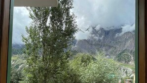 Vista desde una ventana con balcón, con paisaje montañoso, árboles y un edificio a lo lejos.