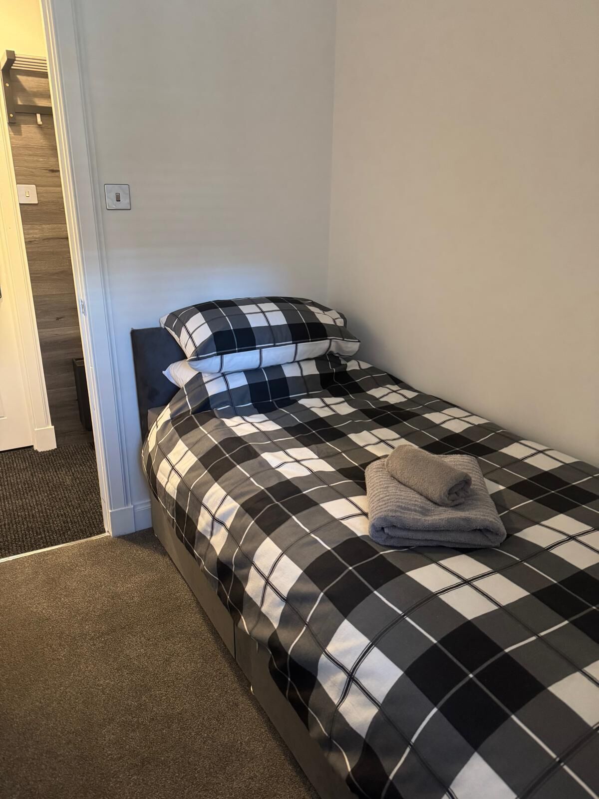 A small, single-bed room with a checkered bedspread and pillows, a folded towel on the bed, and a carpeted floor.