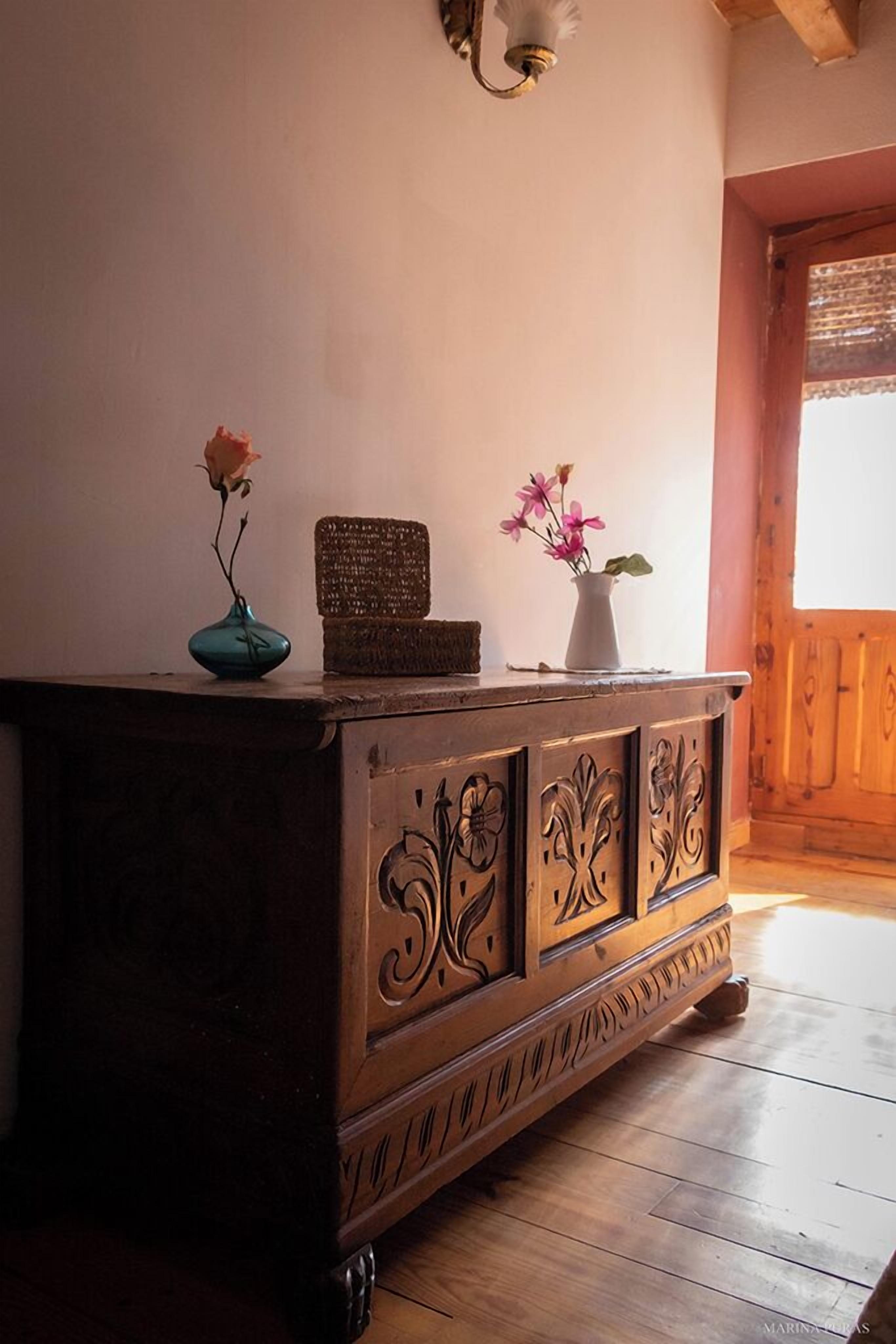 Un gabinete de madera con intrincados grabados, un jarrón azul aguamarina con una sola rosa, y un jarrón blanco con flores rosadas.