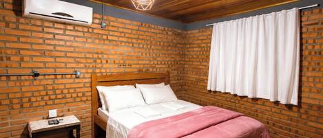 A room with a brick wall, a wooden ceiling, a bed with white bedding, a pink blanket, a bedside table with a lamp, and a window with white curtains.