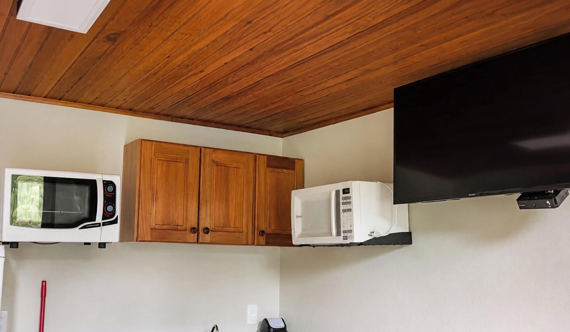 A compact kitchen with wooden cabinets, a microwave, and a flat-screen TV mounted on the wall.