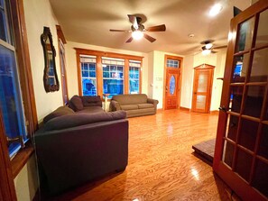 A spacious living room with hardwood floors, two sofas, a ceiling fan, and large windows.