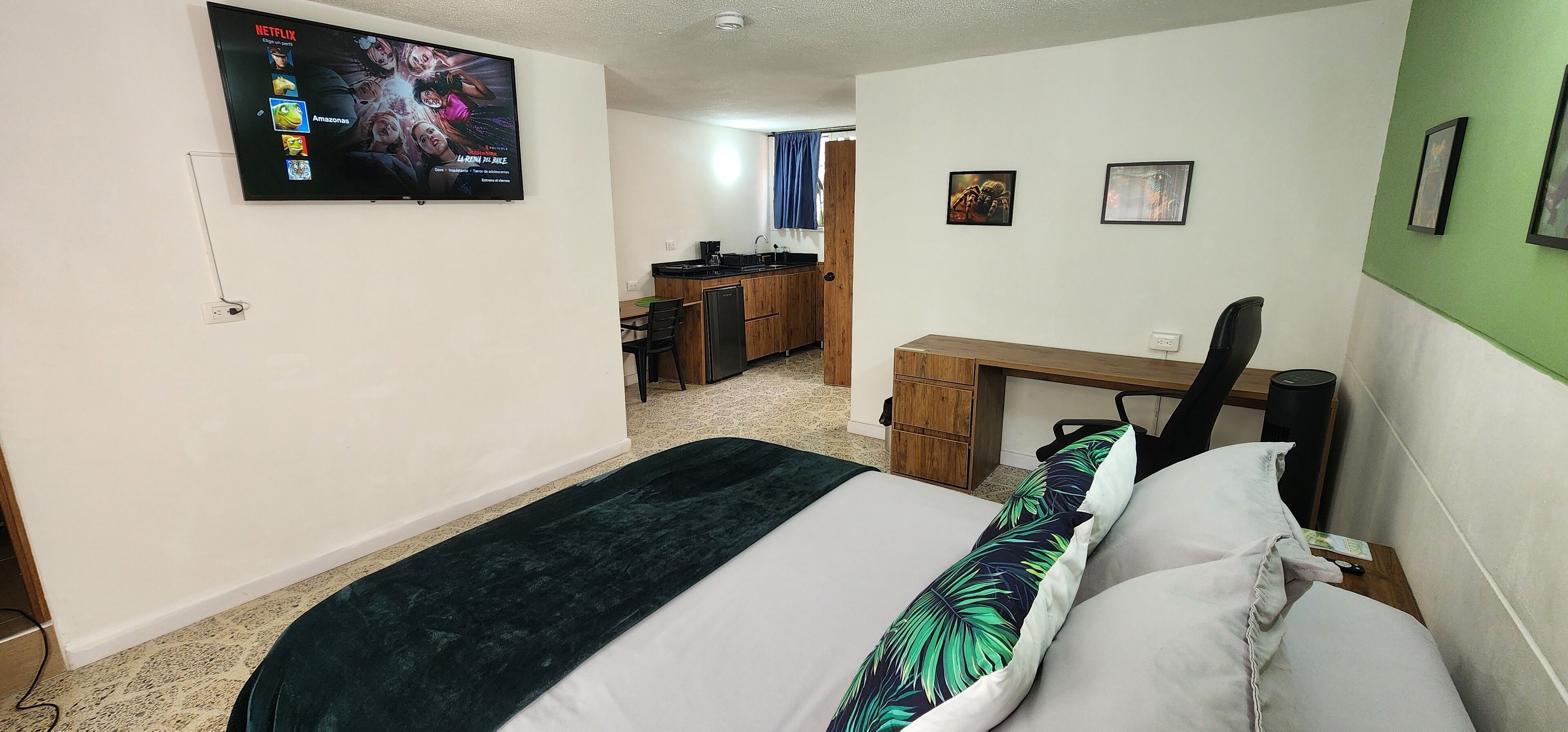 Une chambre d’hôtel avec un lit, un bureau, une chaise, un téléviseur fixé au mur et, au fond, une petite cuisine.