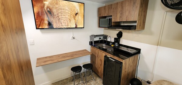 A compact kitchen with a built-in microwave, a coffee machine, and a refrigerator. There are two stools and a wooden shelf.