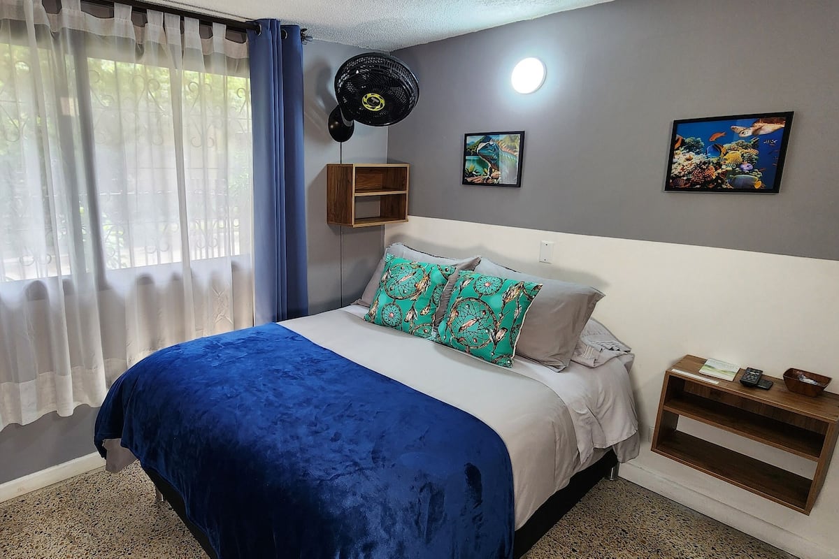 Une chambre à coucher avec un lit, des rideaux bleus, un ventilateur de plafond et des œuvres d’art murales.