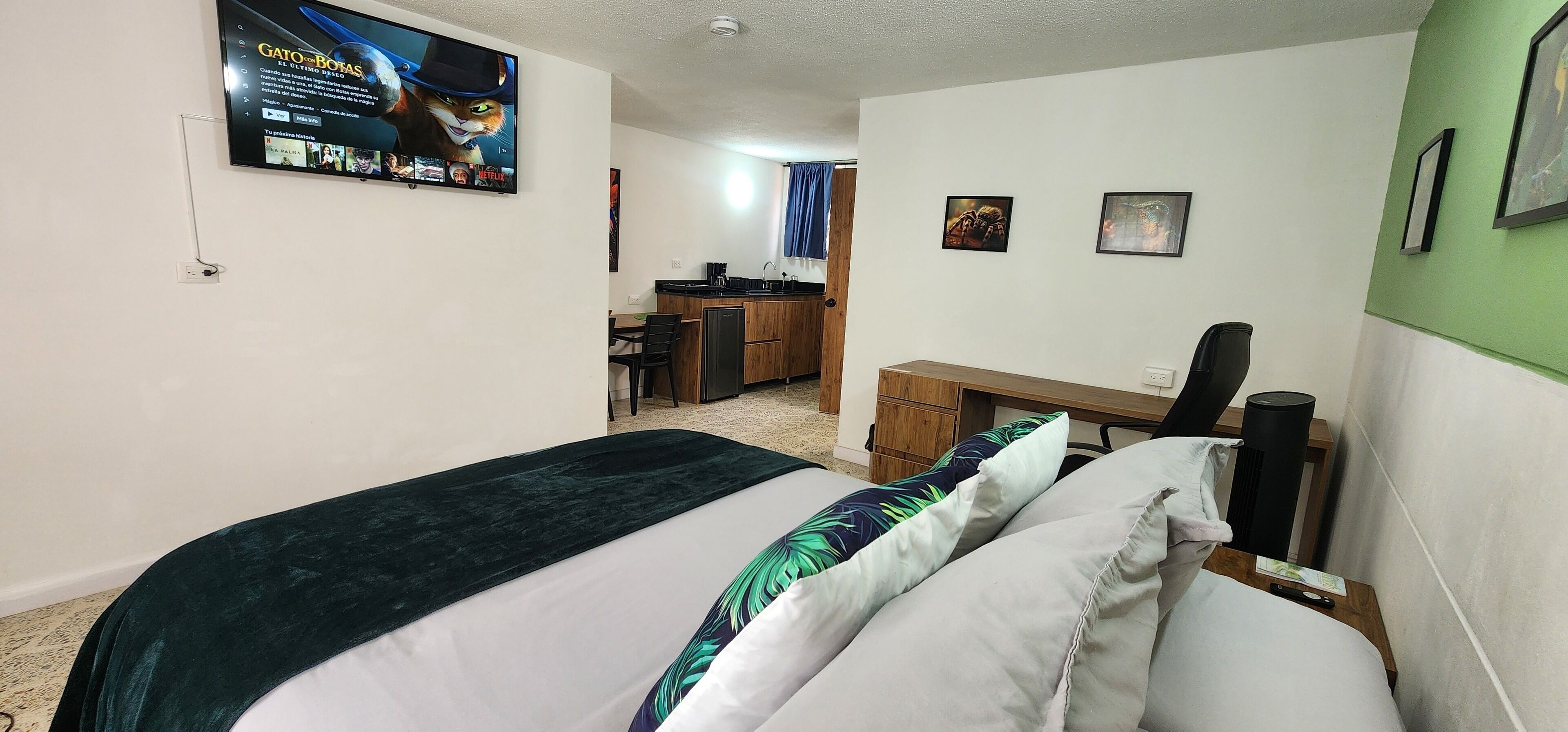 A hotel room with a bed, a desk, a chair, a small kitchenette, and a television on the wall.