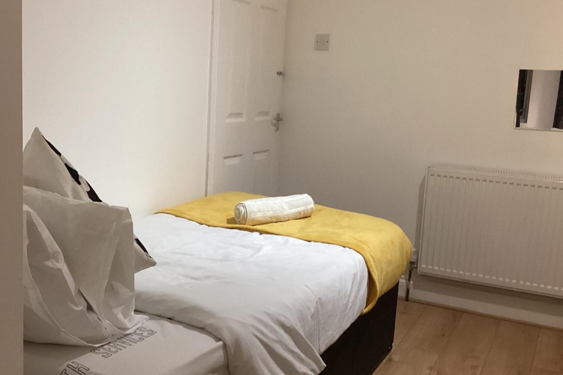 A small, neatly arranged bedroom with a bed, a yellow blanket, and a rolled towel.