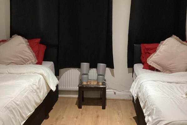 A bedroom with two beds, each with white bedding and red pillows. There is a small wooden table with two gray cups in front of a radiator.