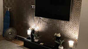 Una sala de estar con un televisor de pantalla plana montado en la pared, una pequeña consola decorativa y un ventilador en el suelo.