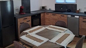 Eine moderne Küche mit Essbereich, ausgestattet mit einem schwarzen Kühlschrank, einem eingebauten Backofen, einem Geschirrspüler und einem großen Fenster mit Jalousien.