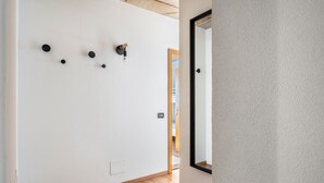 Een gang met een houten vloer, witte muren en een deur met een zwart kozijn.