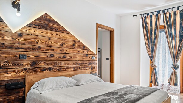 Ein Schlafzimmer mit einem hölzernen Kopfende, einem Bett mit grauer Decke und einem Fenster mit gemusterten Vorhängen.