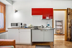 A compact kitchen with white cabinets, a red upper cabinet, a microwave, a kettle, and a sink.