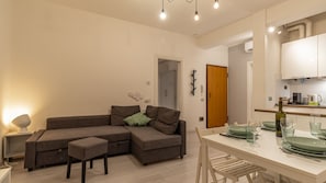A modern living room with a sofa, ottoman, and a dining area with a table set for two. - La casa del Vicolo: 2 camere letto, Wi-Fi & A/C (Modena)