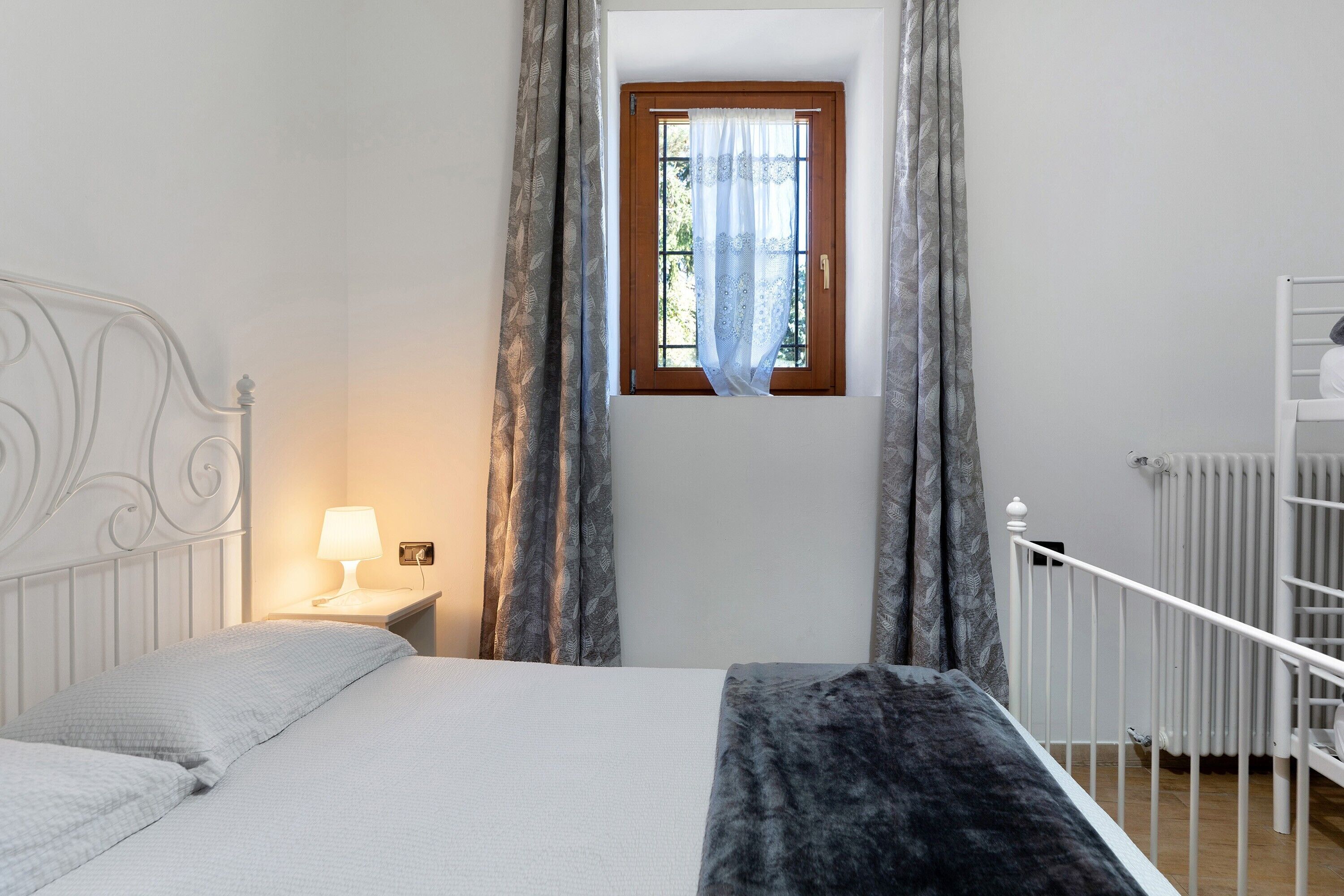 A bedroom with a bed, a nightstand with a lamp, a window with curtains, and a white radiator.