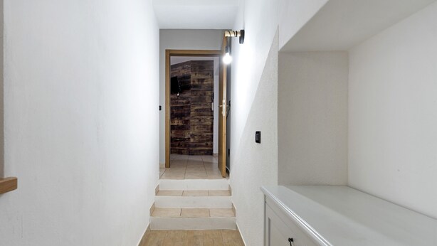 Un pasillo estrecho con una puerta de madera al final, una lámpara en la pared y un armario blanco a la derecha.