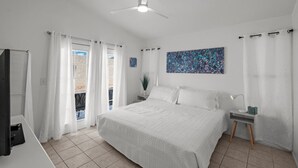 Un dormitorio con cama, mesita de noche, escritorio, ventilador de techo y ventana con cortinas.