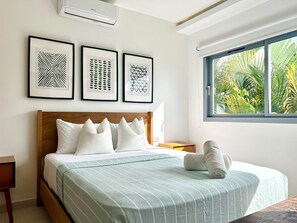 Un dormitorio con cama, almohadas, una ventana que ofrece vista a follaje y tres cuadros en la pared.