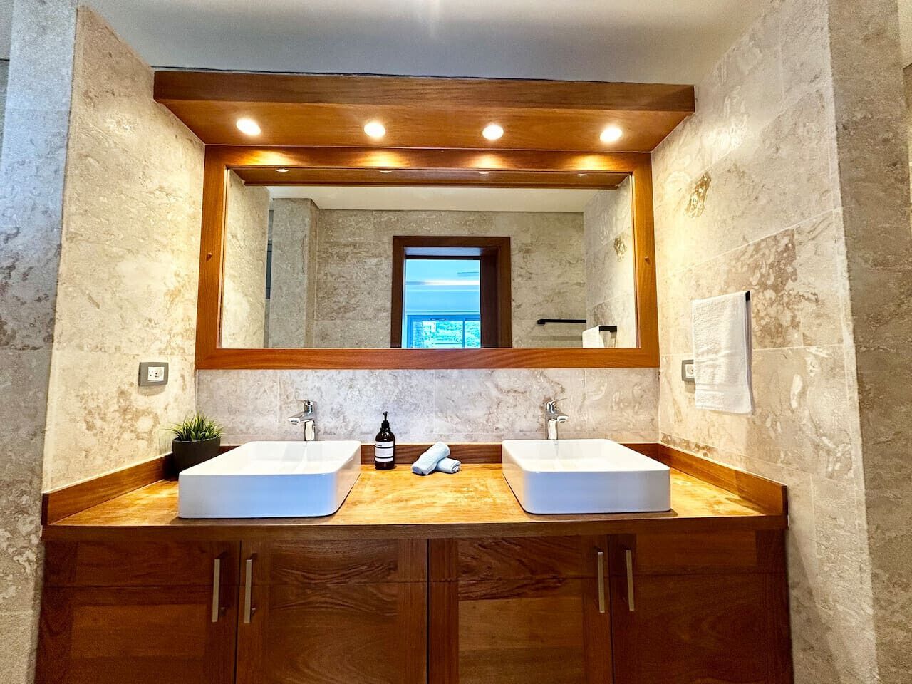 Un baño con dos lavamanos, armarios de madera, un espejo grande y vista al mar a través de una ventana sobre el espejo.