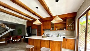 Une cuisine moderne avec des meubles en bois, un îlot central doté de tabourets, un éclairage suspendu et une vue sur l’extérieur grâce à une porte vitrée.