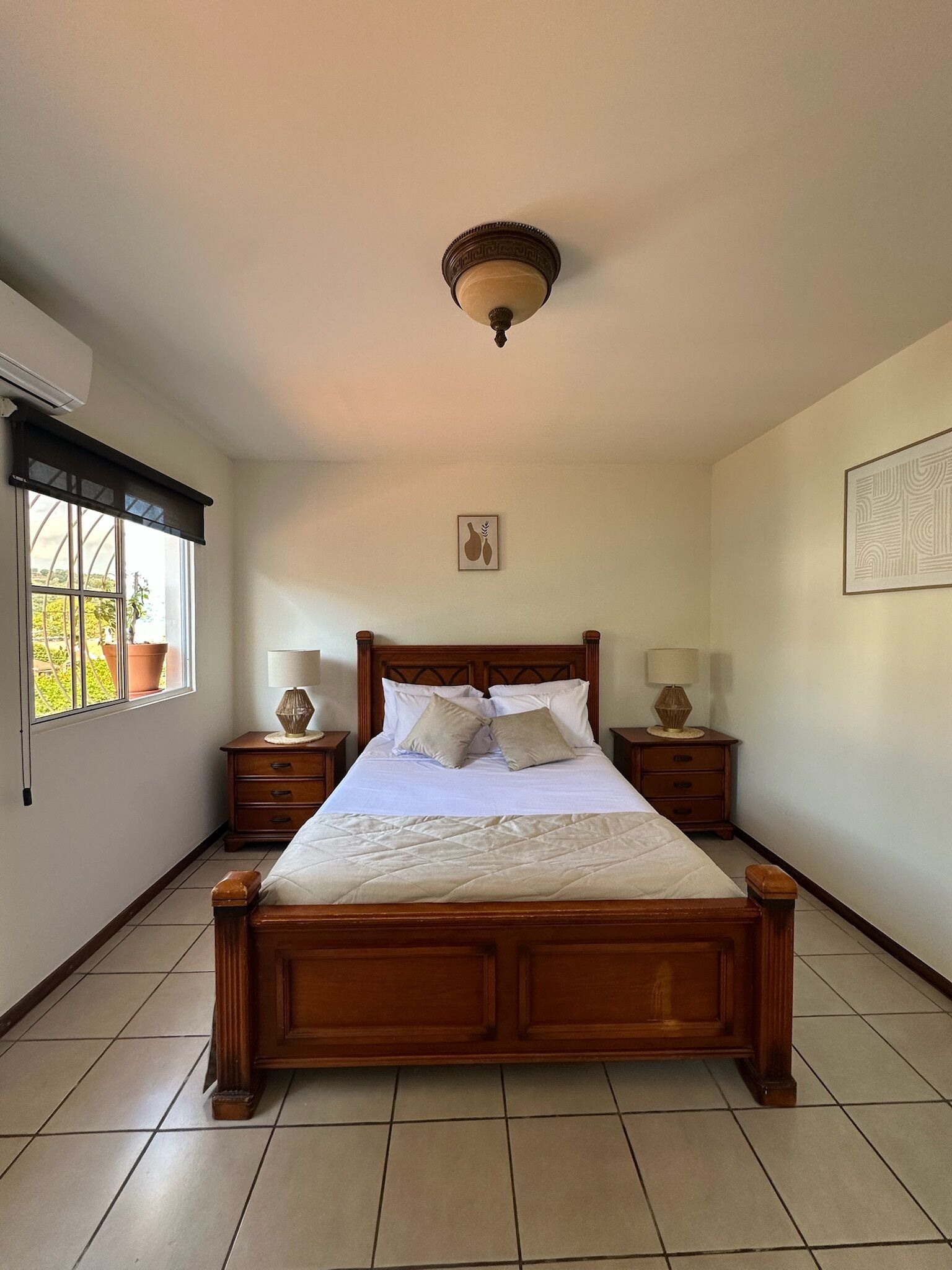 Quarto com cama de madeira, duas mesas de cabeceira com luminárias, ventilador de teto, janela com persianas e um quadro na parede.