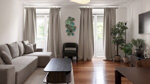 Una sala de estar moderna con sofá, una mesa de centro, una silla y un televisor. Hay cortinas, una obra de arte en la pared y plantas en macetas.