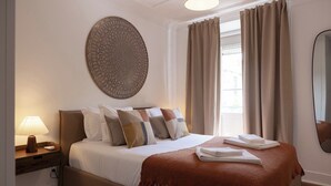 A neatly made bed with a brown throw blanket, a bedside lamp, and a decorative wall piece. - Master Historical Gem in Alfama (Lisboa)