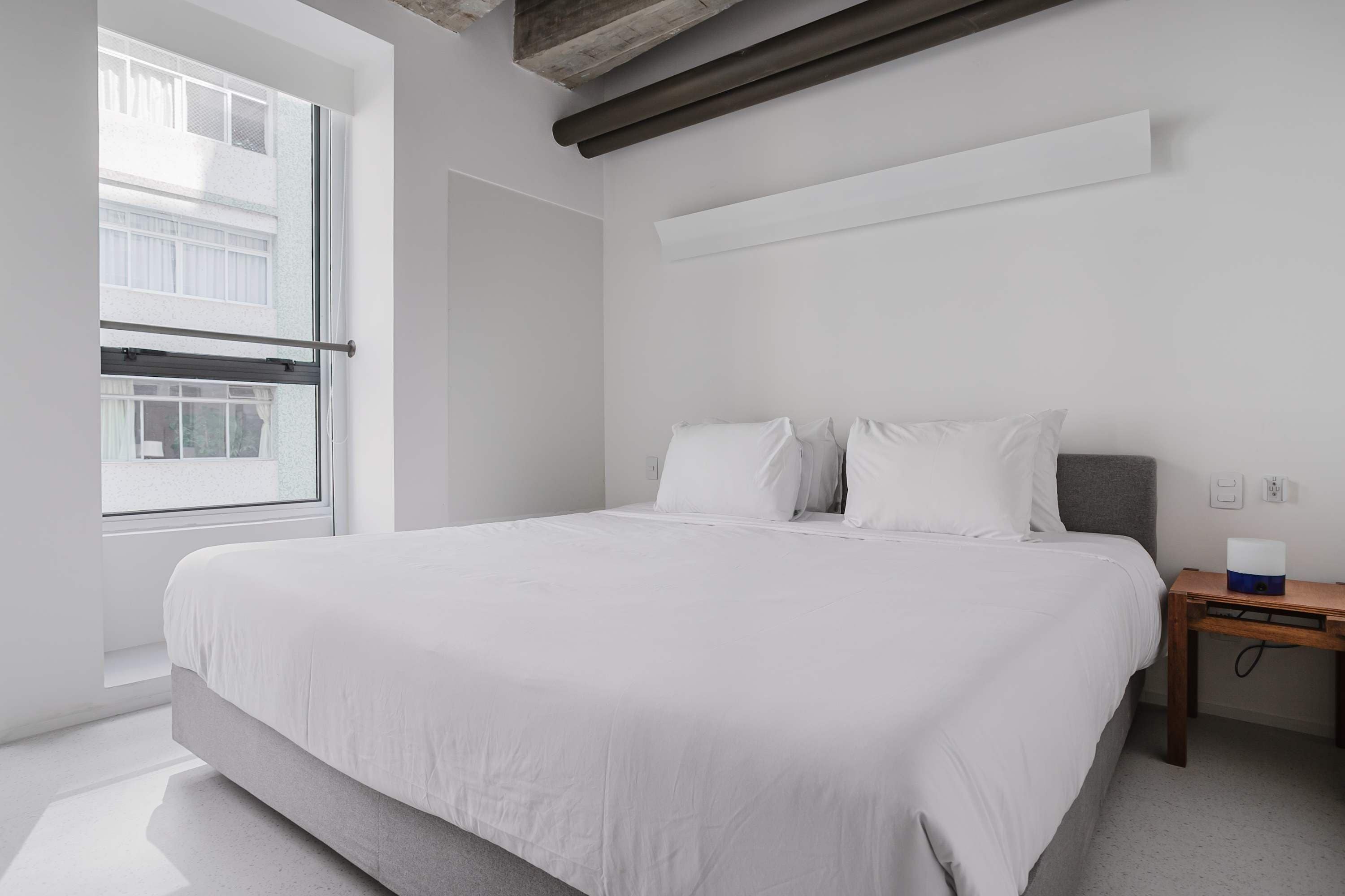 Une chambre à coucher moderne avec un grand lit, une table de chevet en bois et une fenêtre donnant sur des immeubles.