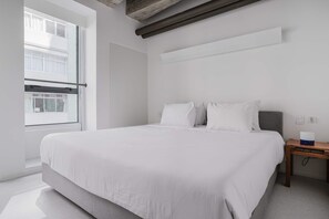 A modern bedroom with a large bed, a wooden nightstand, and a window with a view of buildings.