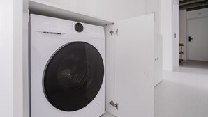 A modern laundry room with a front-loading washing machine, a door with a metal handle, and a visible ceiling with exposed beams. - Eugenio Retreat Stylish 2 BR in Sao Paulo (São Paulo)