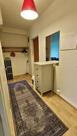 Pieni, hyvin valaistu huone, jossa on puulattia, valkoinen laatikkokaappi, seinälle ripustettu naulakko, punainen riippuvalaisin ja matto.