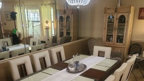 Un espace repas comprenant une grande table en bois, des chaises rembourrées de couleur blanche, un lustre et un buffet aux portes en verre.