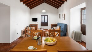 Un'area pranzo con un tavolo di legno, sedie, una ciotola di frutta, una caffettiera e un televisore appeso al muro.