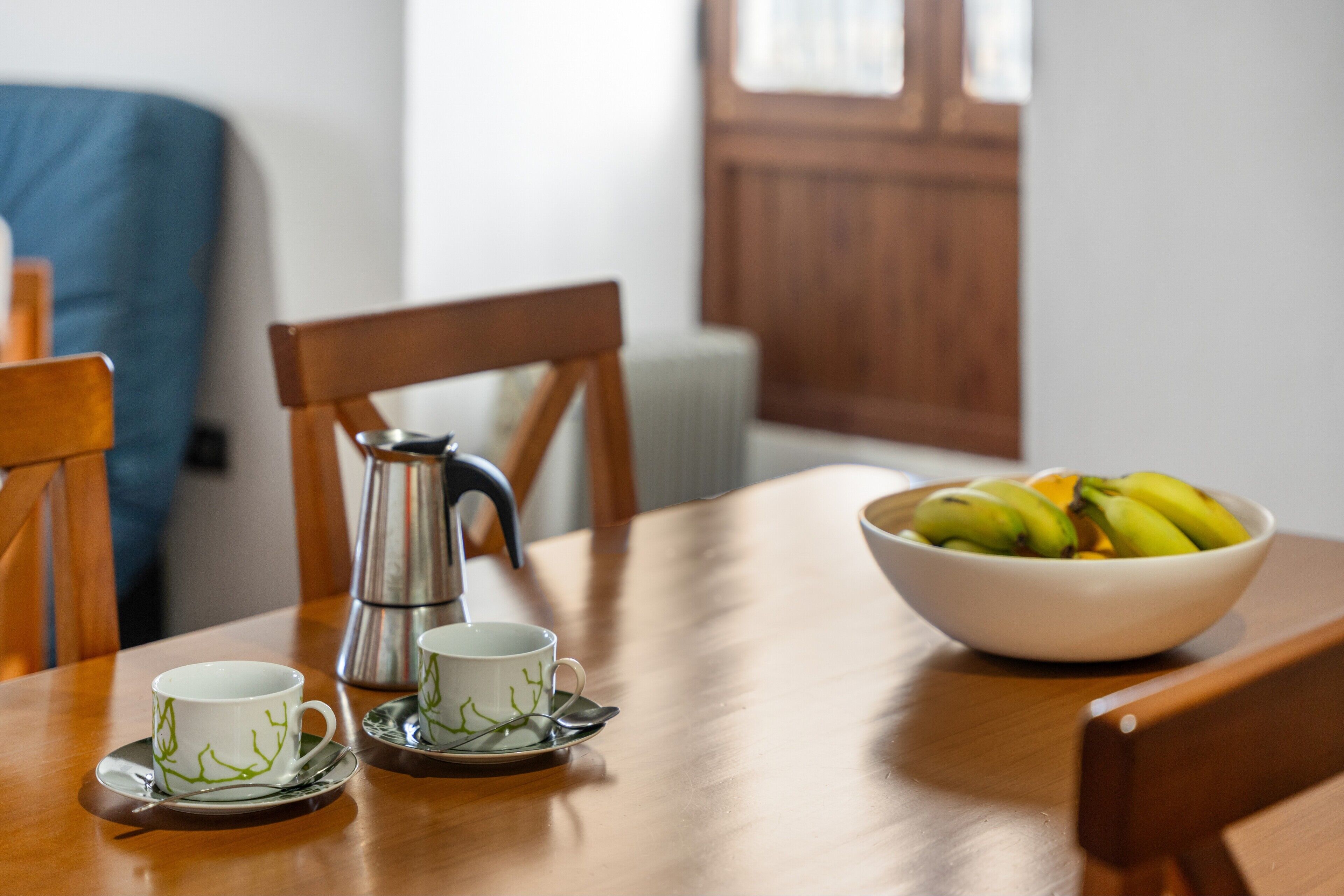 Et træbord med en hvid skål fyldt med bananer, to hvide kopper med grønne motiver og en sølvfarvet kaffekande.