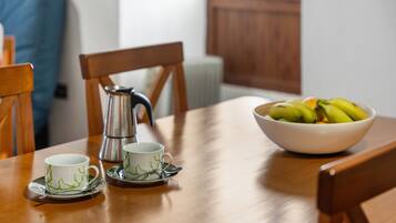 Et træbord med en hvid skål fyldt med bananer, to hvide kopper med grønne motiver og en sølvfarvet kaffekande.