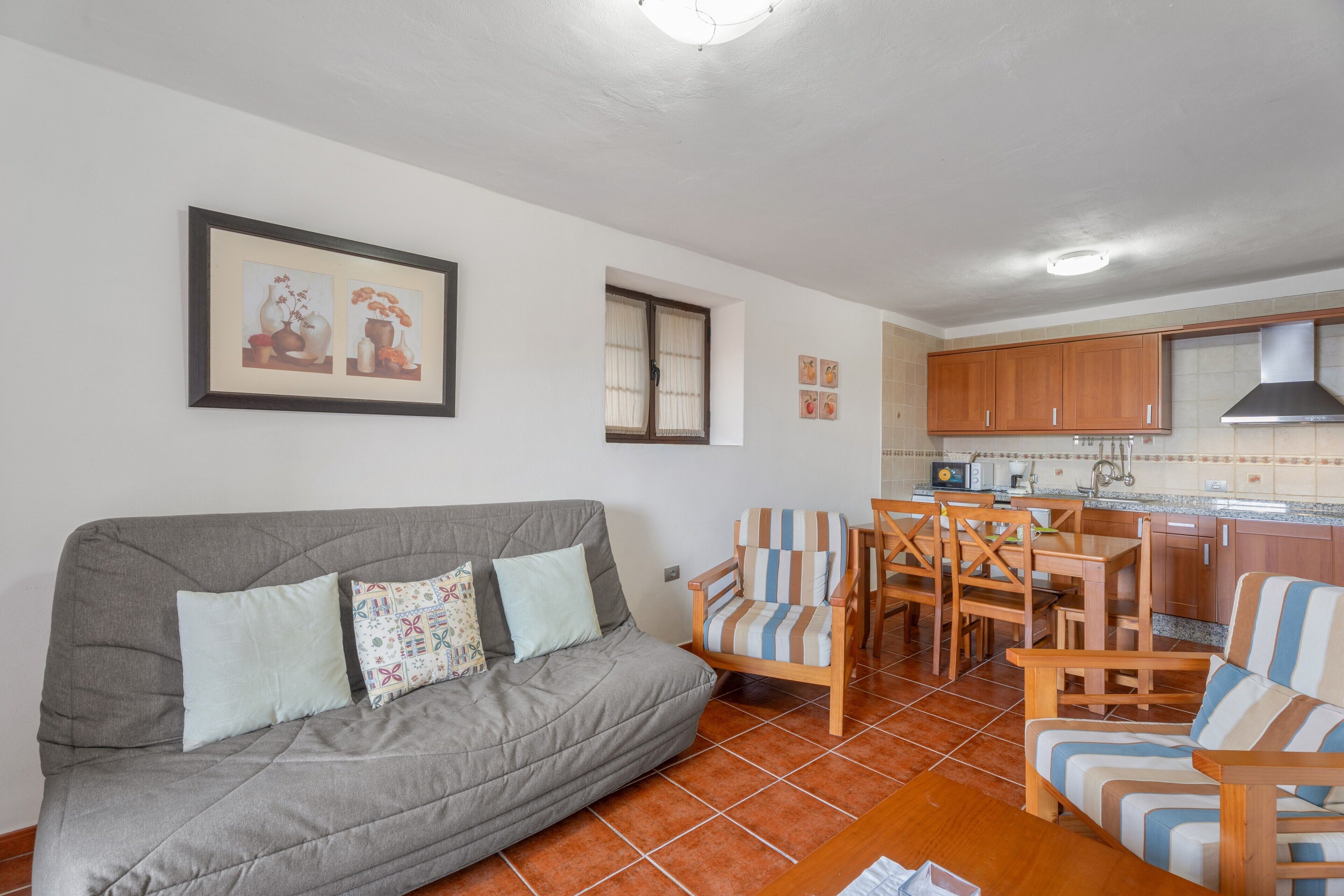 A living area with a sofa, chairs, a dining table, a kitchen with wooden cabinets, and a framed picture on the wall.