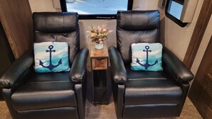 Dos sillones de cuero negro con almohadones a juego, una mesita de madera con un florero, y vistas al exterior a través de una ventana.