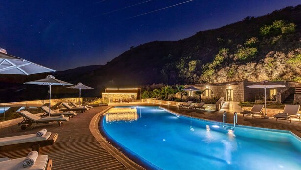 Zona de piscina iluminada durante la noche, con sillones de descanso y sombrillas, rodeada de arquitectura de piedra y montañas al fondo.