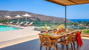 Una terraza con piscina, sillones de descanso y una mesa preparada para comer, con vistas a un paisaje montañoso y al mar.